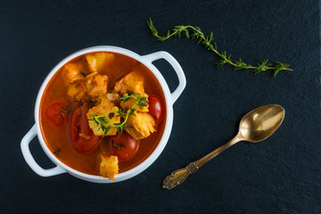Fish soup with cod, tomato, onion, garlic and thyme in white bowl on black stone background, top view.