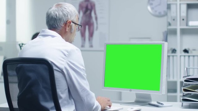 Senior Medical Doctor Working on a Mockup Green Screen Personal Computer. His Assistant Works in the Background. Office has a Touch of Modern Minimalism.  Shot on RED Cinema Camera in 4K (UHD). 
