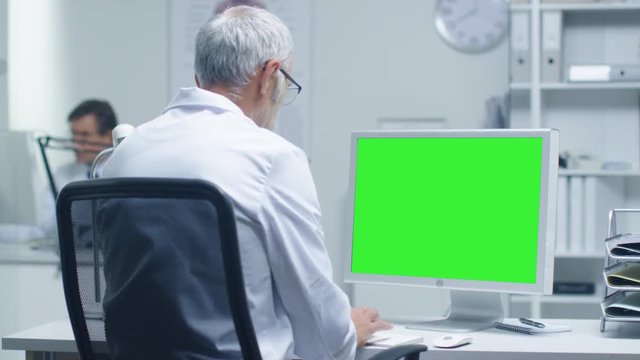 Senior Medical Doctor Working on a Mockup Green Screen Personal Computer. His Assistant Works in the Background. Office is Modern and Representable.  Shot on RED Cinema Camera in 4K (UHD). 