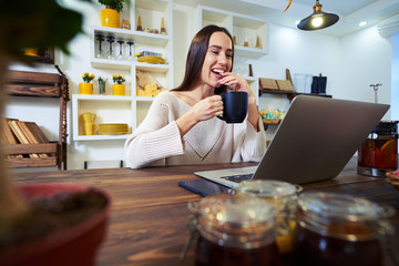 Laughing young woman working on laptop while holding a cup of te