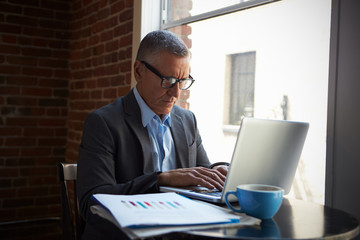 Obraz premium Mature Businessman Working On Laptop By Office Window