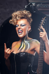 Fototapeta na wymiar Impressed woman punk showing metal horns. Rocker girl in courage posing at camera with her bass guitar, bright makeup and hairstyle. Subculture, lifestyle, art, music, drive concept