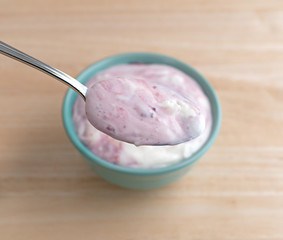 Raspberry yogurt on a spoon with bowl underneath atop a wood table.