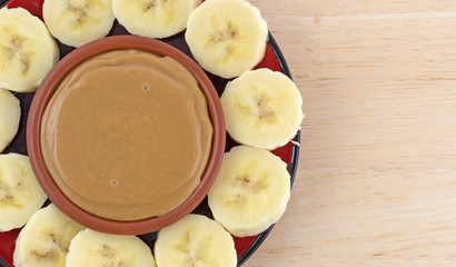 Ripe banana slices in a circle around peanut butter in a small dish on a plate atop a wood table.