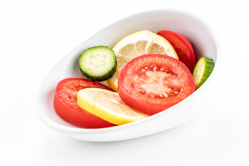 One slanted white bowl with circular yellow lemon red tomato and cucumber slices on a white background