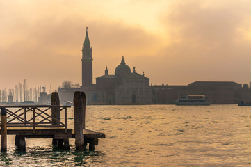 Foggy sunset scene in Venice: old wooden pier and the silhouette of the San Giorgio Maggiore Island behind.