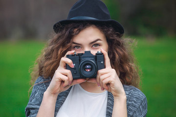 Beautiful girl-photographer with curly hair holds a camera and make a photo, spring outdoors in the park. The concept of tourism and travel.