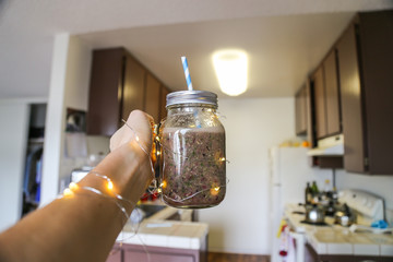 Girl is holding green and red smoothies in glass jar at the kitchen. Warm white lights