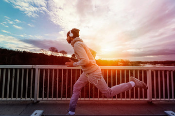 A young man with headphones running on the bridge along a river. Warm tones.