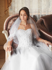 Fototapeta premium Elegant young bride in white wedding dress and veil sitting in ornate armchair, anticipating marriage ceremony