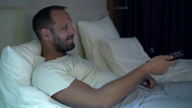 Young, happy man watching TV lying on bed at home at night 
