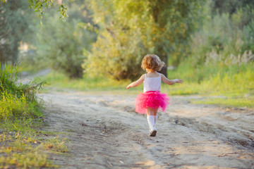 Little curly girl in a bright pink skirt tutu runs on a forest road, the view from the back.