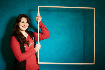 brunette woman in a red blouse standing against the backdrop of a blue wall and holding a large wooden frame