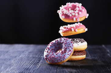 Donut with pink icing and marshmallow in motion falling on a tasty donuts with blue, chocolate and vanilla icing on a dark wooden background.