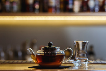 An underexposed lifestyle horizontal image of a teapot with hot alcoholic cocktail on a black tea basis. Wooden bar stand of a restaurant.
