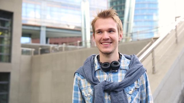 Young man portrait in the city. Caucasian man looking at camera and smiling. He has headphones and pullover on the neck. Lifestyle and city life concepts