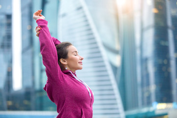 Young sporty girl wearing pink sportswear stretching arms near skyscrapers, preparing for working...