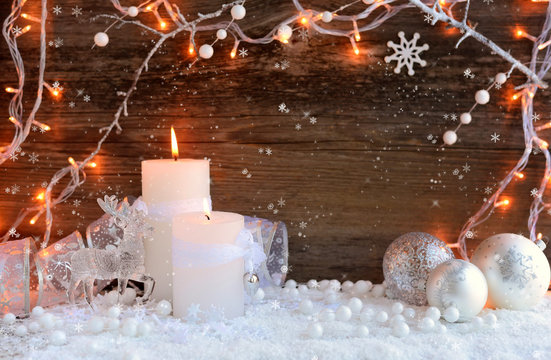 Two burning candles with a deer with christmas decorative balls on snow and Christmas lights. Christmas decorations on wooden background. Festive Christmas background