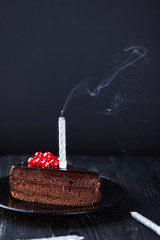 A slice of chocolate cake with redcurrant fork and a single extinguished candle on a dark...