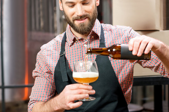 Handsome brewer in apron and shirt pouring beer into the glass at the manufacturing. Image focused on the brewer