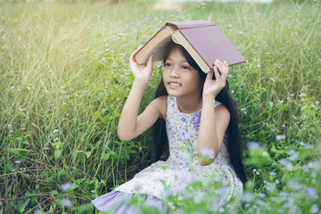 Cute asian little girl with book on head at meadow in holiday