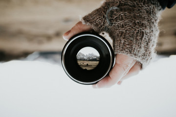 A mountain range photographed through a camera lens held by a human hand with gloves. The picture...