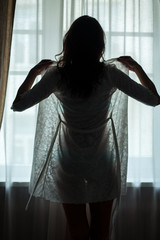 Lady standing near window. Young woman wearing nightie. Beauty is awake.
