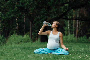 pregnant yoga woman drinking water from a bottle, in the lotus position. park ,grass ,.outdoor, forest.