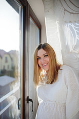 Charming pregnant woman stands near a window with white curtains.