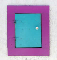 Small handmade aqua door with raspberry colored frame set into white stucco wall. Vertical.