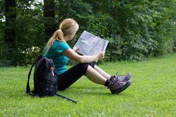 A young woman sits in grass and looks into the tourist map, backpack and walking sticks put aside