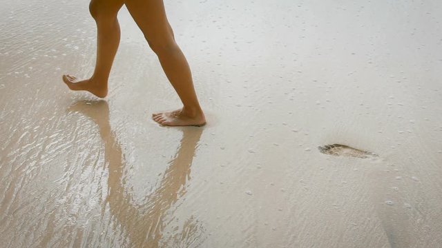 Beautiful girl in a black bathing suit goes on wet sandy beach and leave a trail. Young woman with slim, tanned legs walking along the shoreline.