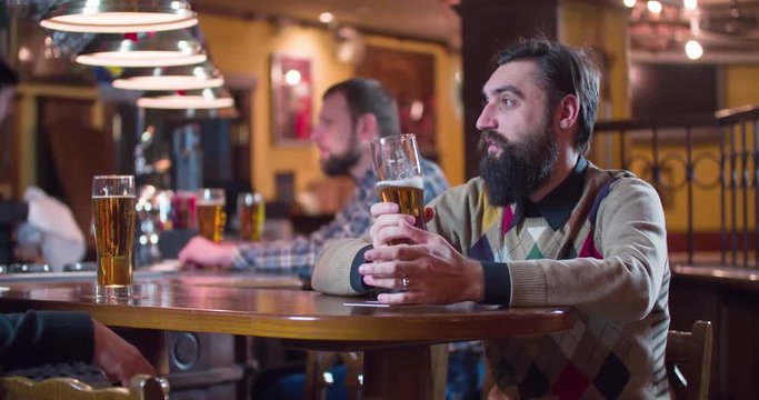Male friends talks and drinks beer at pub bar 4k video. Men taste lager from glasses, bartender and other guests at the background. 