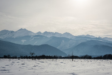 Scenic winter view with the mountains covered with snow on the side of the road from a plain field on a background of clouds and sunny sky