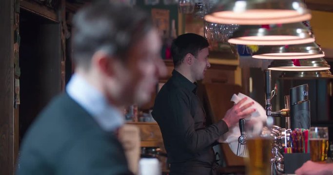 Speaking smiling man at beer pub bar counter close-up 4k video. Working bartender in background with refocus