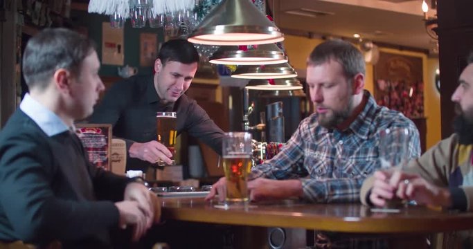 Bartender bringing beer glass for client at pub bar 4k video. Men friends drinks lager, talks, enjoy and laugh. High five with barman