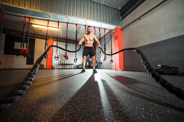 Young fit man exercising with battle ropes in the fitness gym