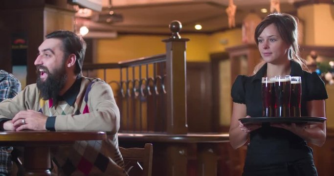 Waiter brings beer glasses for men 4k video on pub, bar or restaurant. Male friends doing toast, cheers and having fun. Man looks at girl tits
