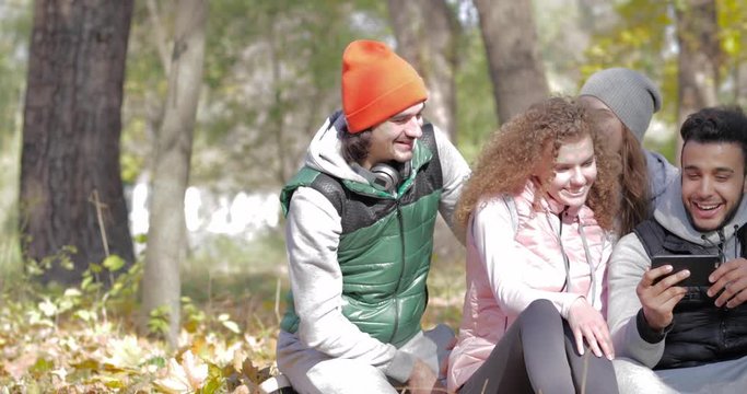 Young People Group Sit On Ground In Forest Watch Funny Video On Smart Phone Outdoor, Friends Laughing Autumn Park Slow Motion 60 Fps