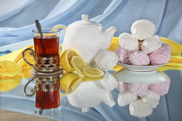 Still life with tea, lemon, white and pink marshmallows. Black tea in a glass with a cup holder, lemon, marshmallow on the table with reflection