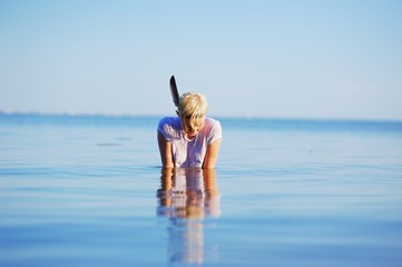 Young pretty blonde girl sitting in the water with his head down in the sun on the blue sea surface, closeup.