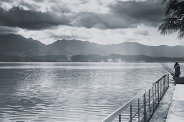 fisherman fishing at quiet lake with mountain sky and cloudy countryside black and white tone.