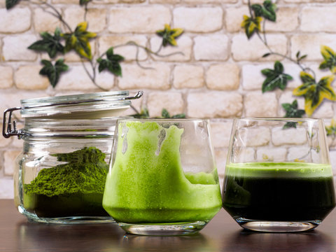 Two glasses of green juice, one being empty,  and a jar of green juice powder
