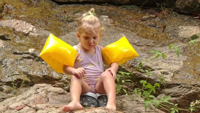 Little Blond Girl in Arm-hands Sits Scratches Leg on Stone