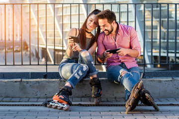 Couple holding cell phones. Two rollerbladers sitting. Sharing new photos. © DenisProduction.com