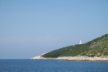 White high coastal lighthouse on a hill with green trees above sea and rocks in Greece.
