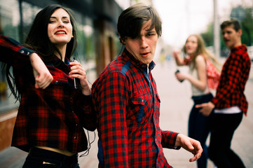 Two happy trendy teenage friends walking and dancing in the city, listening to the music with headphones, talking each other and smiling. Lifestyle, friendship and urban life concepts.