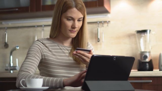 Beautiful attractive young woman purchasing online with tablet by credit card at home in the kitchen