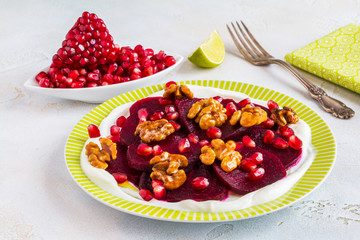 Dietary vegetarian salad of roasted beets with pomegranate seeds, walnuts caramelized in honey and natural yoghurt. Slices of lime, vintage fork, white and gray table.