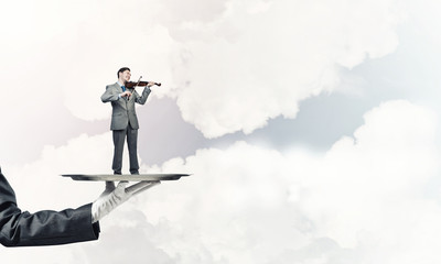 Businessman on metal tray playing violin against blue sky background